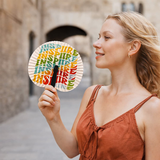A person holds a multicoloured paper fan featuring the word "INSPIRE" repeatedly, with a black handle.
