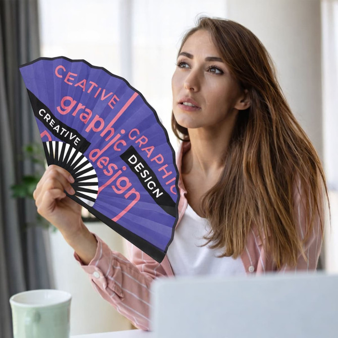 A bamboo fabric hand fan in purple and black, featuring graphic design elements, held by a woman near a laptop.