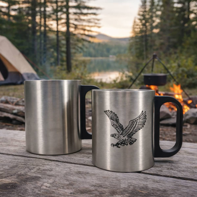 A stainless steel insulated mug with a plastic handle, featuring a design of an eagle. Set outdoors by a campfire.