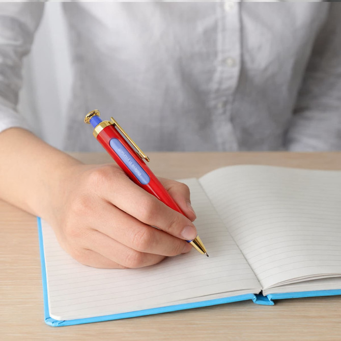 A red and blue pen with a gold tip is held over an open notebook on a wooden table. The pen has a logo.