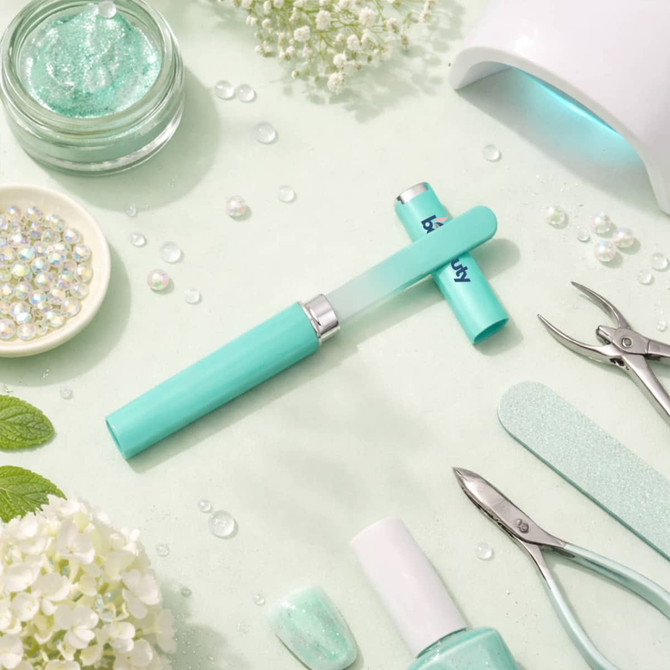 A gradient glass nail file in turquoise, surrounded by nail care tools, glitter, and decorative elements on a pale green background.
