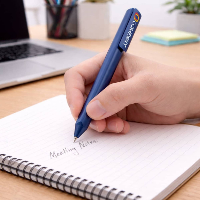 A blue triangular grip pen is held above a notepad with handwritten notes on a wooden desk with a laptop in the background.