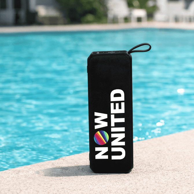 A black portable speaker with a round logo, sitting by a blue pool.
