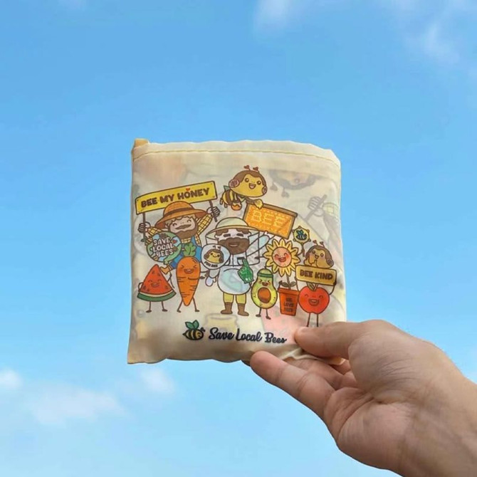 A foldable shopping bag in soft yellow with colourful cartoon characters and a logo, set against a blue sky.