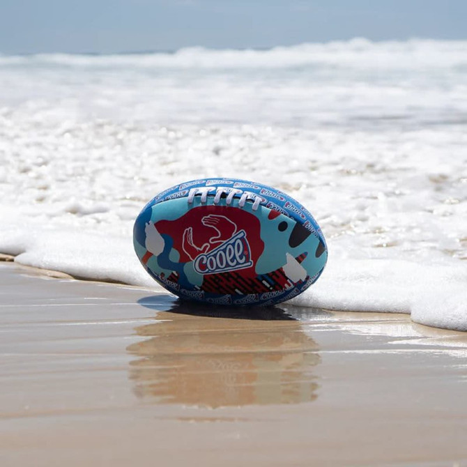 A blue and red soft AFL football rests on wet sand near the ocean, with waves gently rolling in.