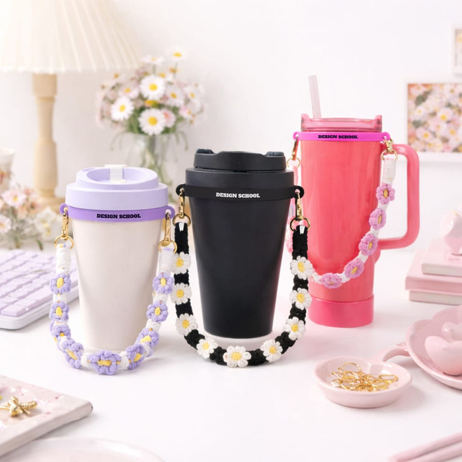A drink bottle with a woven flower strap in black, pink, and purple, displayed on a pastel-themed table.