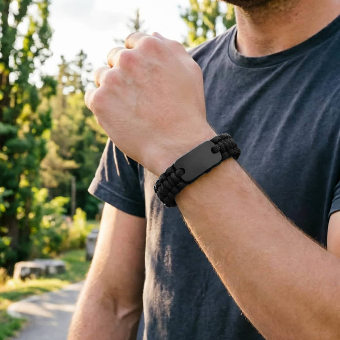 A black paracord bracelet featuring a metal plate, worn on a wrist amidst a natural outdoor setting.