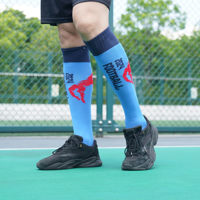Knee high sports socks in blue with red pattern and a logo, worn with black athletic shoes on a green court.