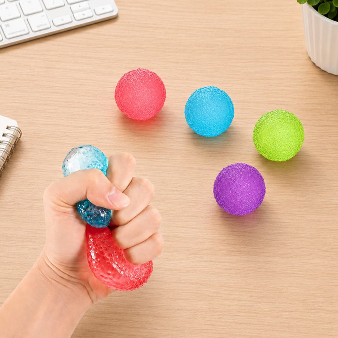 A hand squeezing a clear squishy stress relief mini ball, surrounded by coloured stress balls in red, blue, green, and purple.