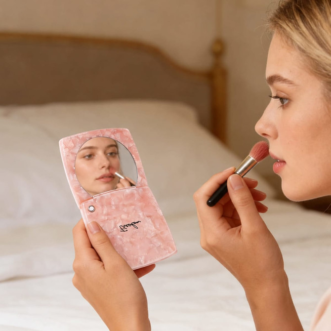 A pink acetate vanity mirror with a compact design, held by a person applying makeup in a softly lit setting.