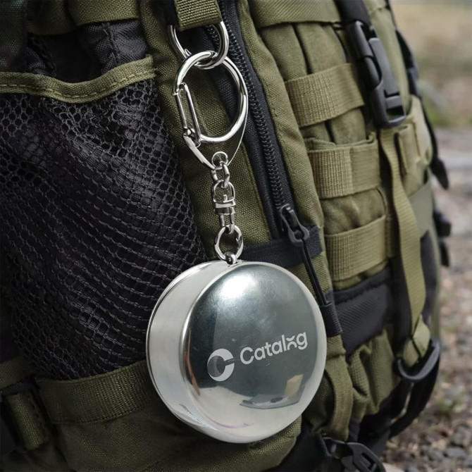 A compact, stainless steel folding cup, silver in colour, attached to an olive green backpack with a carabiner.