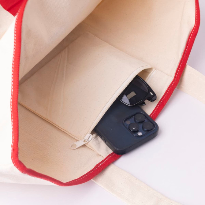 A red and cream canvas tote bag with a zipped pocket containing sunglasses and a mobile phone.