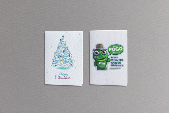A large seed paper greeting card featuring a Christmas tree design and a green character illustration with a logo.