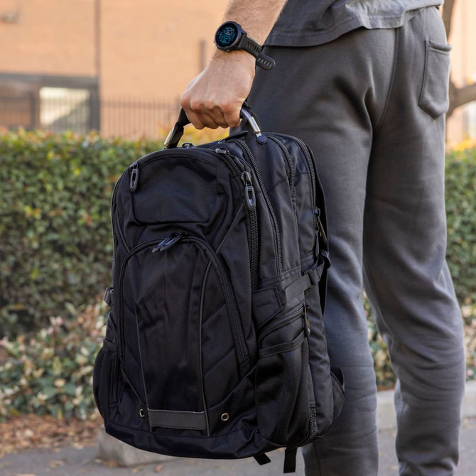 A black Eiger Backpack is held by a person wearing grey pants and a watch, set against a green hedge background.