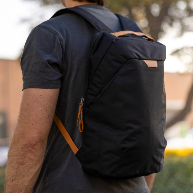 A person wearing a black backpack with an orange accent, featuring a logo, positioned against a blurred background.