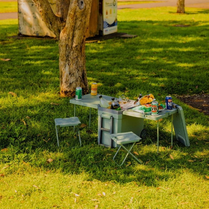 A green cooler box with an attached table and two chairs, set on grass with drinks and snacks around it.