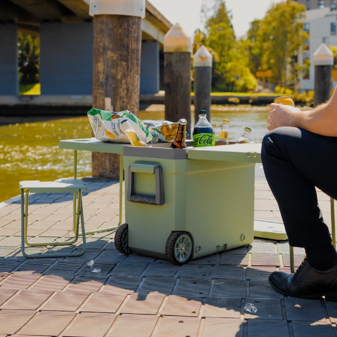 A green cooler box with wheels, featuring a table and chair, set by a waterway with snacks and drinks on it.