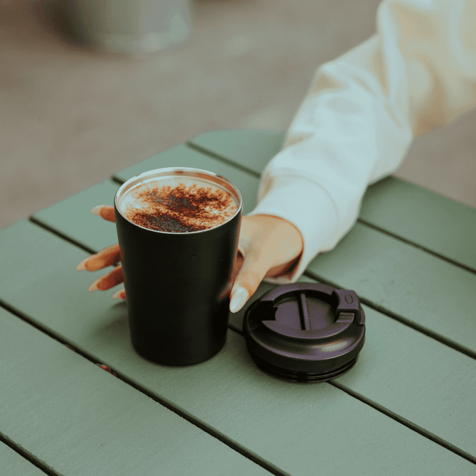 A double-walled 360ml coffee cup in black with a purple lid resting on a table beside a hand.
