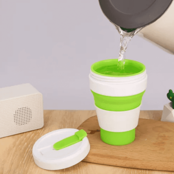A collapsible silicone cup in green and white, being filled with water, beside a speaker on a wooden surface.