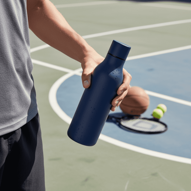 A drink bottle in navy blue held by a person, with sports equipment in the background.