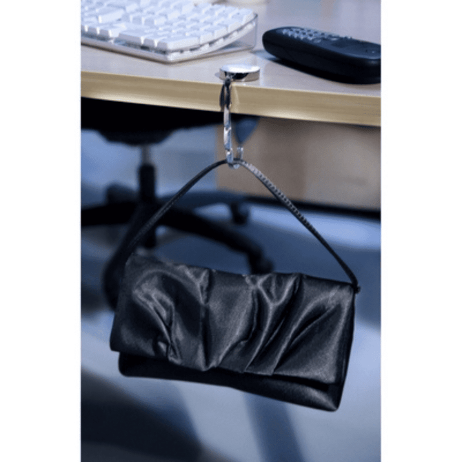 A black bag hanger clips onto a desk, supporting a black clutch. A computer keyboard and remote are in the background.