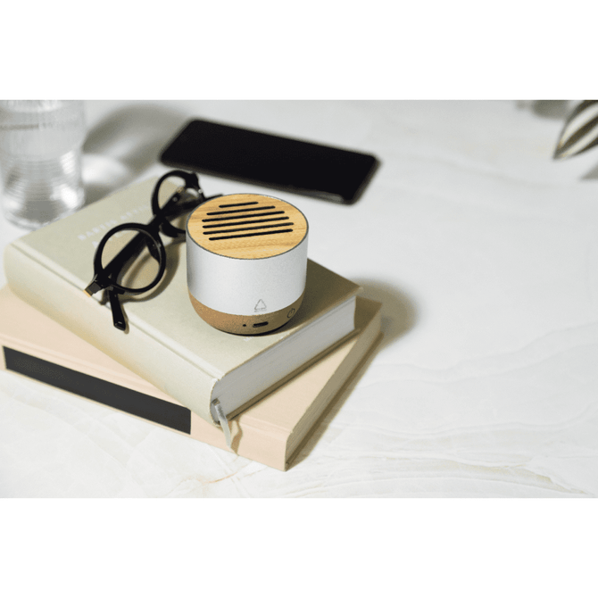 A compact white and wood wireless speaker sits on a stack of books, alongside glasses and a smartphone.