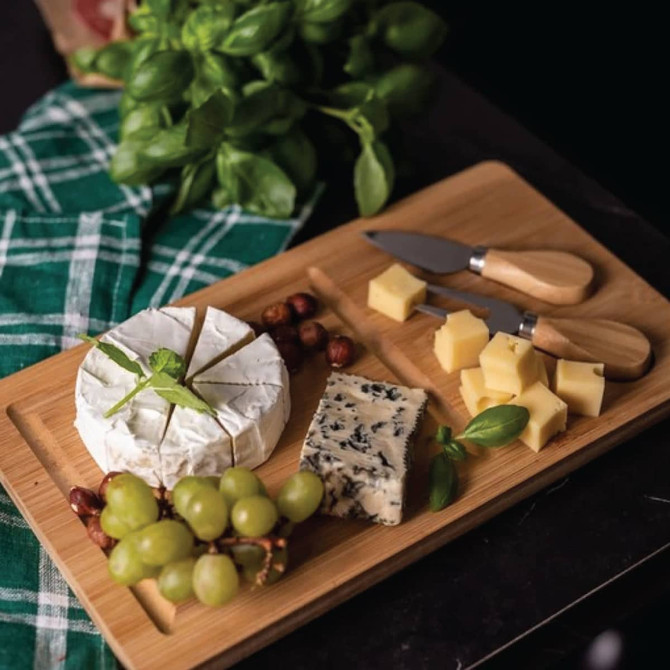 A bamboo cheese board with a circular cheese, assorted cheeses, grapes, and two cheese knives with wooden handles.