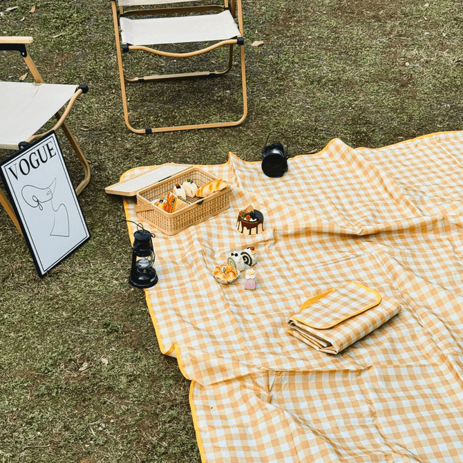 A yellow gingham picnic blanket spread on grass, surrounded by picnic items, a chair, and a framed artwork.