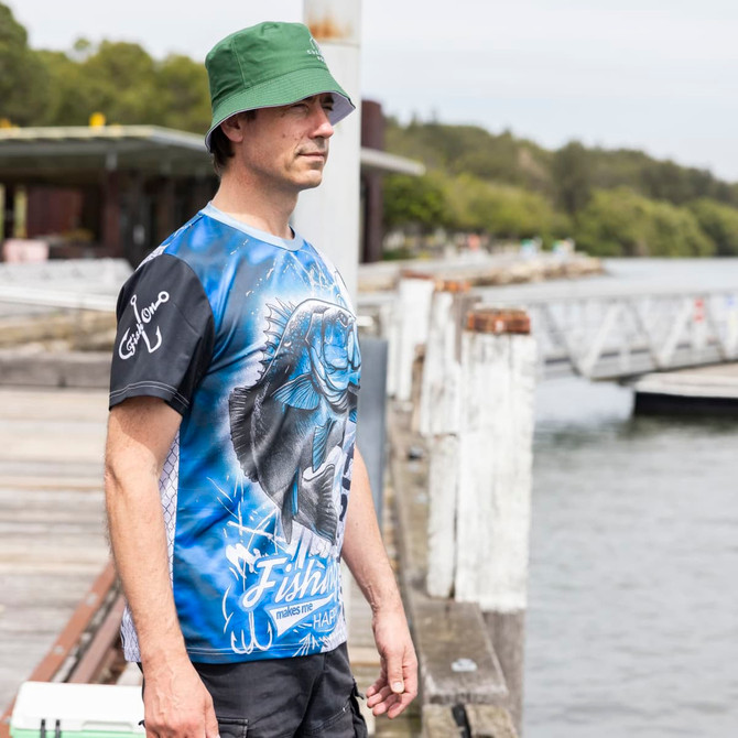 A man wearing a blue and black polyester sublimated fishing tee and a green bucket hat by the water.