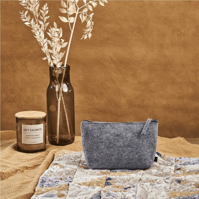 A grey RPET felt mini cosmetic bag is positioned on a textured surface, alongside a candle and a vase with dried foliage.