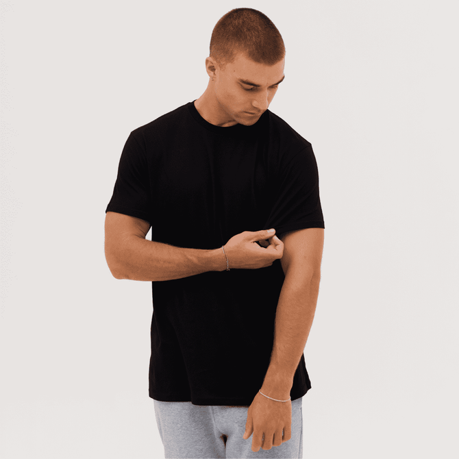 A black T-shirt on a male model adjusting the sleeve, with a plain background.