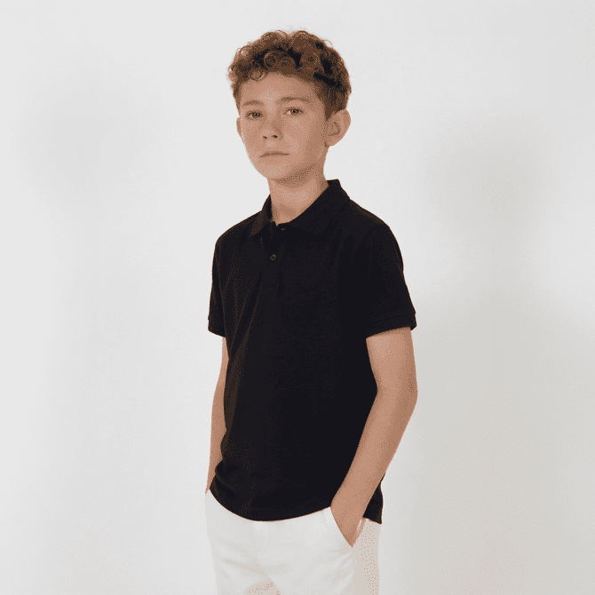 A youth black polo shirt with a collar, featuring short sleeves and a pocket. The model is standing with hands in pockets.