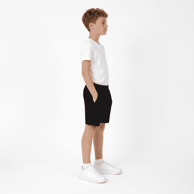 A pair of black youth shorts featured on a child wearing a white t-shirt and white shoes, standing in profile.