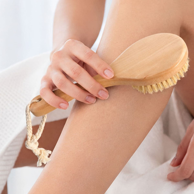 A round body brush with a short wooden handle and natural bristles, being used on skin, with a rope loop for hanging.