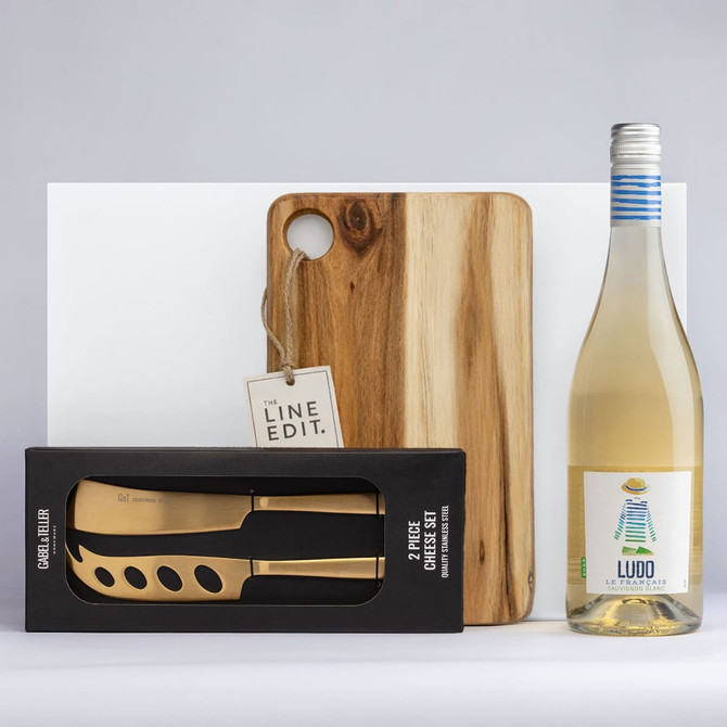 A wooden cheese board, a two-piece gold knife set in a black box, and a bottle of white wine with a colourful label.