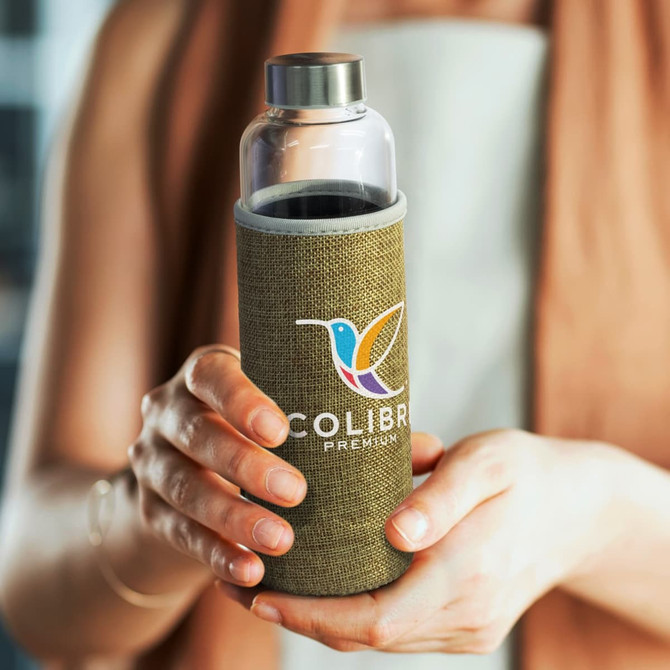 A drink bottle with a clear glass body and a green natural sleeve, held by a person with an outstretched hand.