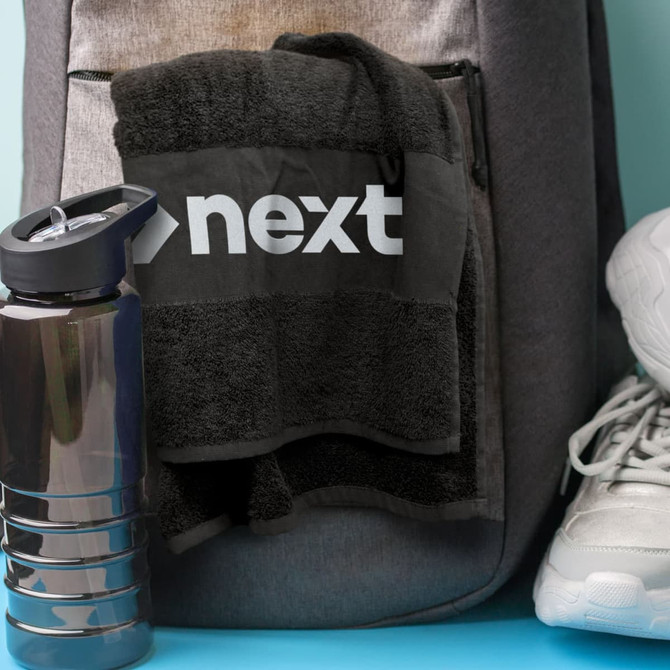 A black gym towel with a logo hangs from a grey backpack, next to a drink bottle and white running shoes.