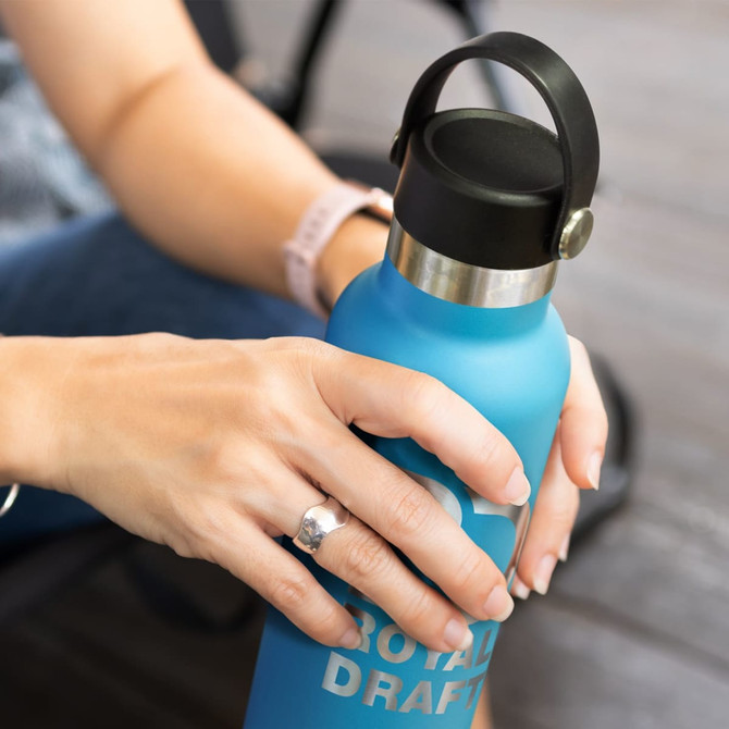 A drink bottle in blue with a black carry lid, held in a person's hands. The bottle features a branding logo.