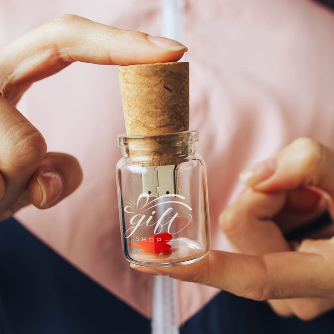 A glass bottle with a cork stopper contains a flash drive. The bottle has a logo and is held between two fingers.
