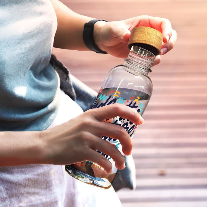 A drink bottle made of clear material, featuring a wood lid and colourful designs, held by a person.
