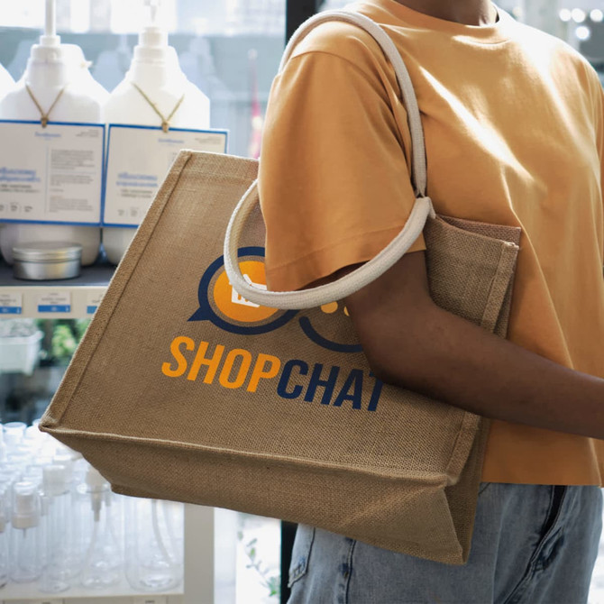 A jute tote bag in natural tones with a logo and sturdy handles, being carried by a person in a casual outfit.