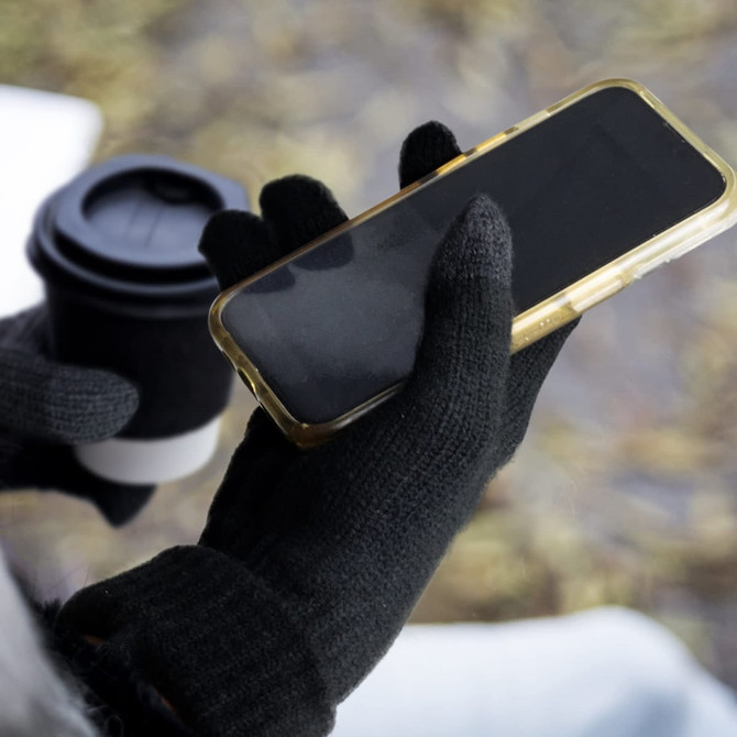Black Himalayan Tech Gloves holding a smartphone, with a coffee cup partially visible in the background.