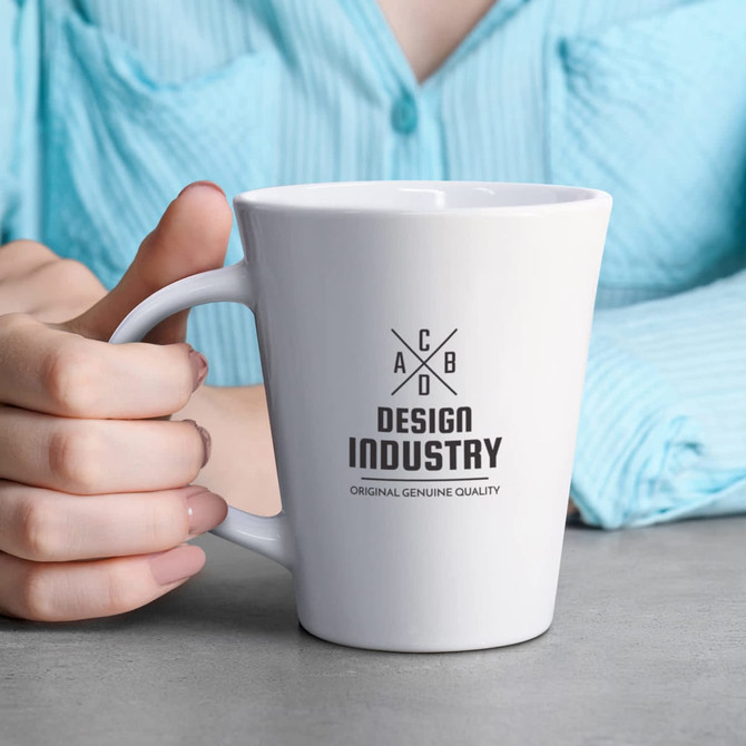 A white coffee mug with a logo, held in a hand, on a grey surface, with a blue shirt in the background.
