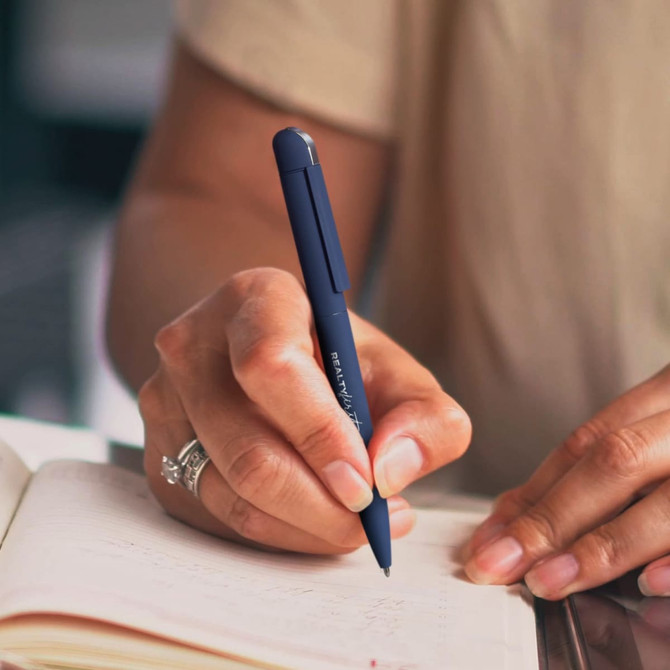 A hand holding a navy blue metal pen, writing in a notebook. The pen has a sleek design and features a logo.