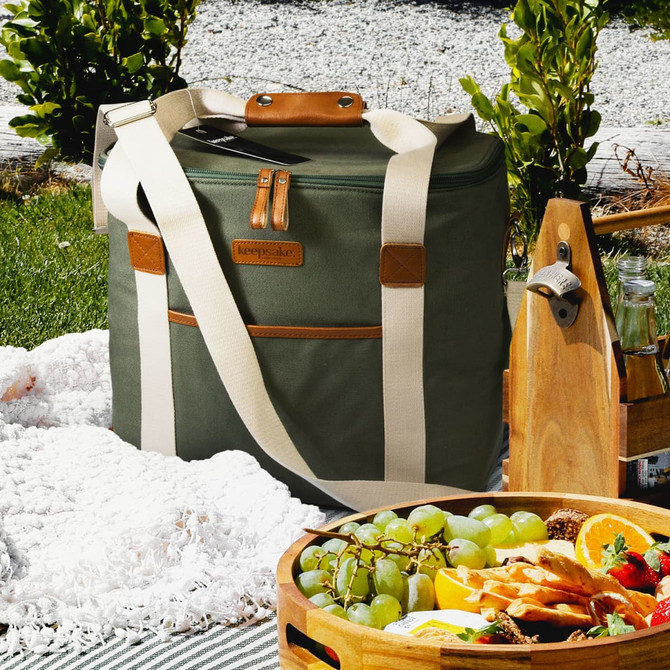 A green cooler bag with a tan leather strap and logo, resting on a picnic blanket beside a fruit platter.