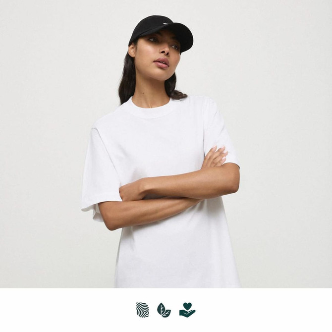 A white t-shirt displayed on a person wearing a black cap. The shirt has a relaxed fit and includes branding.