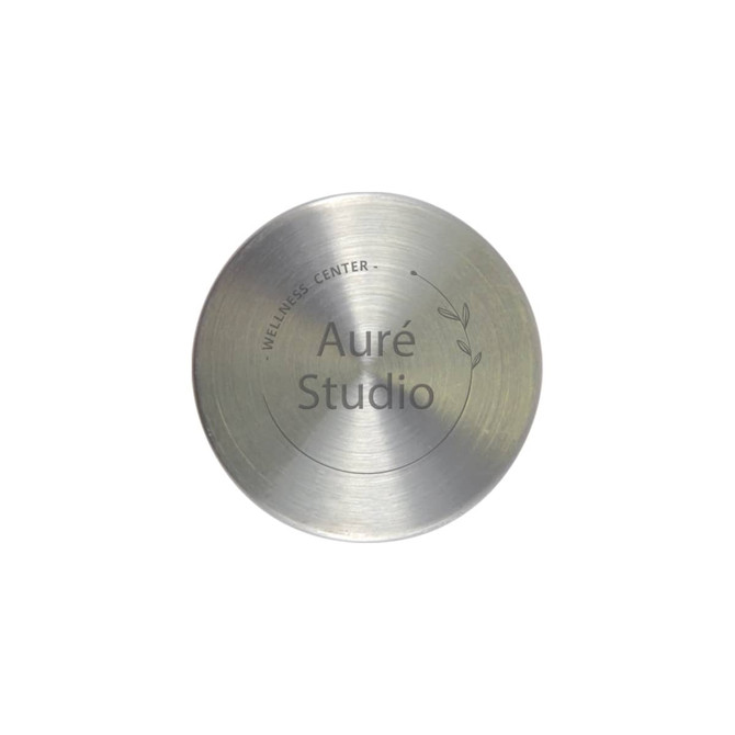 A stainless steel circular plate with a minimalist design featuring the words "Auré Studio" and a decorative leaf motif.