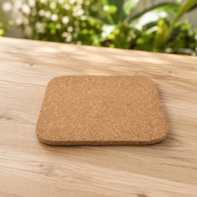 A cork square coaster with rounded edges, set on a wooden table with greenery in the background.