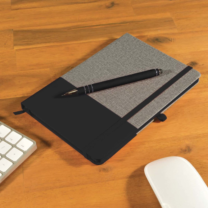 A gray notebook with a black cover and a black pen resting on top, placed on a wooden desk alongside a keyboard and mouse.