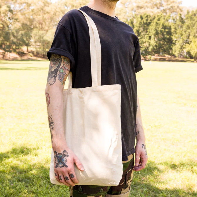 A beige tote bag with long handles is held by a person standing outdoors on grass. The bag has a logo.